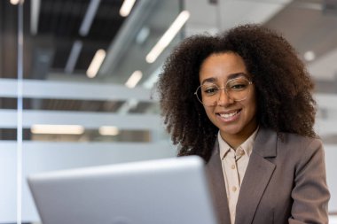 Ofiste dizüstü bilgisayar kullanarak gülümseyen Afro-Amerikan iş kadını. Kendine güvenen, gözlüklü ve takım elbiseli genç bir kadın masasında özenle çalışıyor. Modern işyeri ve başarı kavramı.