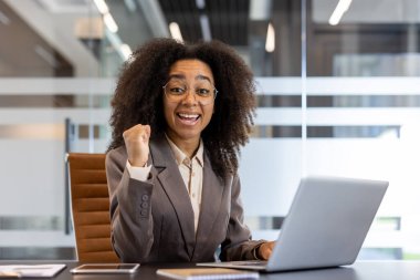 Genç Afrikalı Amerikalı iş kadını heyecanlanıyor ve iş yerinde başarıyı kutluyor. Gözlüklü ve takım elbiseli gülümseyen kadın modern ofisin içinde dizüstü bilgisayarla çalışıyor. Pozitif duygular ve profesyonel
