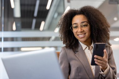 İş yerinde gülümseyen ve dizüstü bilgisayar kullanan genç Afrikalı Amerikalı iş kadını. Modern ofiste kendine güvenen bir profesyonel. İş iletişimi, üretkenlik, teknoloji kavramı.