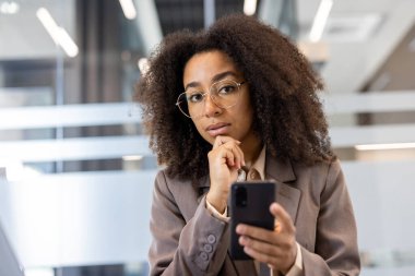 Genç Afro-Amerikalı iş kadını ofiste telefonla iş kıyafeti giyiyor. İş konusunda çalışan ve düşünen kendine güvenen profesyonel bir yönetici. Modern işyeri ortamı.