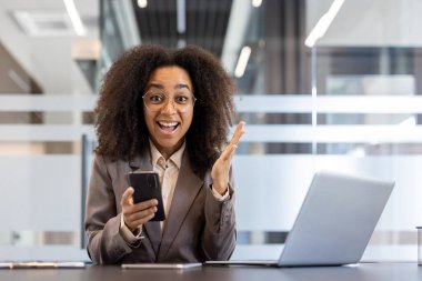 Genç Afrikalı Amerikalı iş kadını iş yerinde telefon kullanırken heyecan gösteriyor. Dizüstü bilgisayarlı profesyonel ortam, modern ofis ortamı.