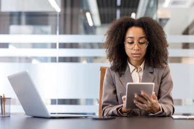 İş yerindeki iş yerinde tablet kullanan genç Afrikalı Amerikalı iş kadını. Tablet ve dizüstü bilgisayarda odaklanmış profesyonel çalışma verimliliği, modern çalışma ortamının gösterimi