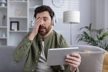 Kulaklık takan bir adam evde kanepede oturmuş hayal kırıklığına uğramış bir ifadeyle tablet kullanıyor. Stres, yorgunluk ve teknoloji sorunları.