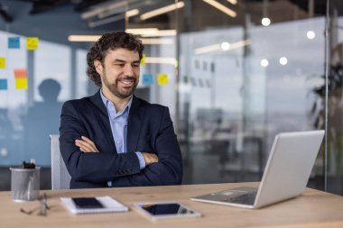 Modern ofiste dizüstü bilgisayara bakarken kendine güvenen işadamı gülümsüyor. Masasında tablet, defter ve telefonla oturan takım elbiseli bir profesyonel..