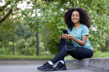 Afro-Amerikalı bayan sporcu açık hava fitness dersine ara veriyor kulaklıkla müzik dinlerken gülümsüyor. Yeşil park ayarları rahatlamayı vurguluyor