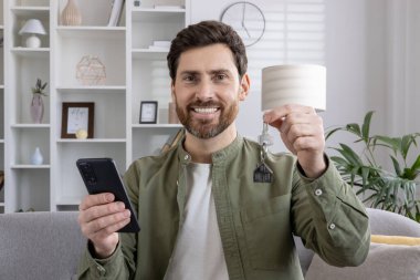 Yeni evin anahtarını elinde tutan gülümseyen adam, kapalı alanda akıllı telefon kullanıyor. Gayrimenkul alımını kutlayan mutlu bir ev sahibinin portresi. Başarı kavramı, ev sahipliği, teknoloji.