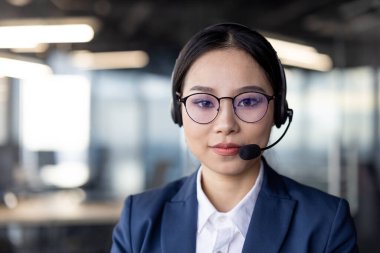 Profesyonel Asyalı bir iş kadınının portresi, gözlük ve kulaklık takıyor, modern ofis ortamında özgüvenle çalışıyor. Müşteri hizmetleri ve çağrı merkezi işlemlerini tasvir etmek için ideal