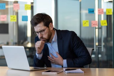 Ofiste dizüstü bilgisayar başında çalışırken öksüren ve göğsünü tutan bir iş adamı. Sağlık sorunu yaşayan, rahatsızlık ve endişe gösteren, muhtemelen hastalık ya da solunum sorununu gösteren kişi.