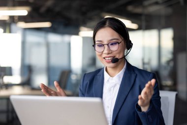 Kendine güvenen Asyalı iş kadını kulaklık takıyor ve sanal bir toplantı sırasında konuşuyor. Modern bir ofis alanında oturuyor, bir dizüstü bilgisayar kullanarak dinleyicileriyle aktif olarak iletişim kuruyor..