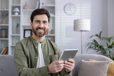 Evde tablet kullanırken kanepeye oturmuş gülümseyen bir adam. Rahat ve neşeli bir ifade, günlük kıyafetler giyme, ve cihaza bağlanma. Parlak ve rahat oturma odası ortamı