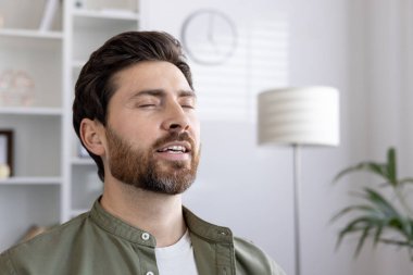 Young man with closed eyes practicing mindfulness meditation and relaxation at home. Concept of stress relief, inner peace, and mental well-being through meditative practice. clipart