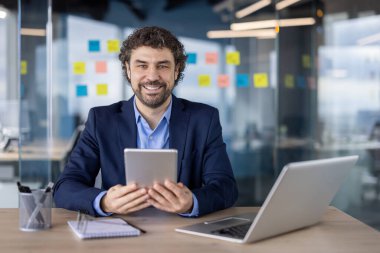 Kendine güvenen işadamı tablet kullanırken gülümsüyor ve akıllı telefon ve dizüstü bilgisayarla masada oturuyor. Profesyonel takım elbiseli, modern ofis ortamında çalışıyor..