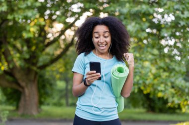 Neşeli Afrikalı Amerikalı kadın başarılı spor salonunun ardından yoga minderini tutuyor ve cep telefonundan olumlu bir mesaj izliyor. Atletizm kıyafeti giymiş.