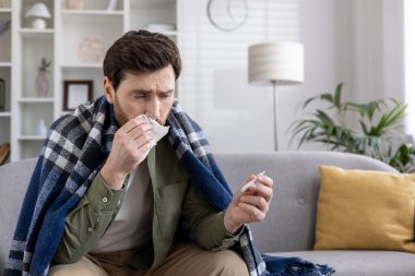 Battaniyeye sarılı kanepede oturan ve elinde mendil ve termometre tutan adam gripten muzdarip. Rahatsızlık ifadesi, kötü hissetmek, sıcaklığı kontrol etmek ve soğuk algınlığı belirtileri. sağlık ve hastalık kavramı.