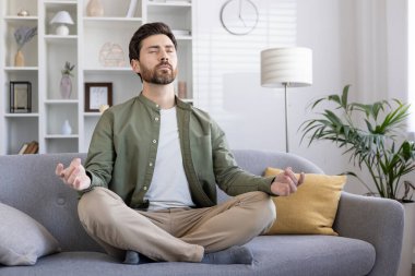 Evdeki kanepede gözleri kapalı meditasyon yapan adam sakin ve sakin bir ifade takınıyor. Konforlu yaşam alanındaki rahatlama, farkındalık, sükunet ve kişisel sağlığın vurgulanması.
