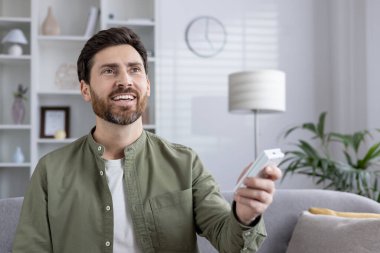 Tatmin olmuş bir adam klimayı uzaktan kumandayla elinde tutuyor, içeride gülümsüyor. Adam sıcaklığı uzaktan kumandayla ayarlıyor. Konfor, teknoloji ve refah kavramı.