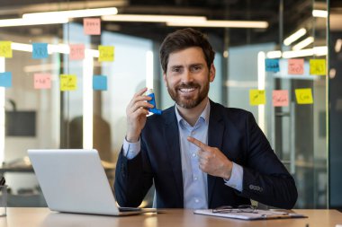 Gülümseyen iş adamı, modern ofiste çalışırken astım ilacı kullanıyor. Takım elbiseli adam dizüstü bilgisayar ve defterin yanındaki astım spreyini işaret ediyor. Pozitif ifade, sağlık konsepti, iş-yaşam dengesi.