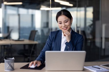 Modern bir ofiste dizüstü bilgisayarla çalışan mavi takım elbiseli Asyalı bir iş kadını. Gülümsüyor ve işleriyle meşgul görünüyor. Kurumsal atmosferi olan profesyonel bir ortam..