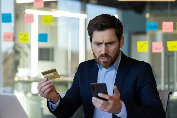 stock image Concerned businessman in suit holding credit card and phone, looking at phone screen with worried expression, dealing with online banking or payment problem in modern office setting