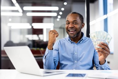 Afro-Amerikalı olgun iş adamı dizüstü bilgisayarda çalışıyor, elinde heyecandan para tutuyor. Finansal başarı, mutluluk ve mesleki başarı kavramlarını yansıtır.