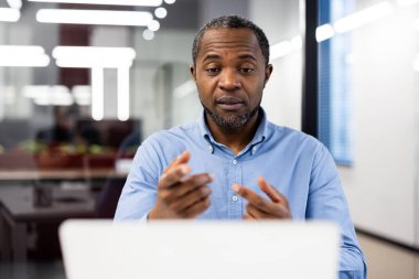 Modern ofis ortamında ciddi bir iş adamı çevrimiçi toplantı yapıyor. Orta yaşlı bir adam, bilgisayar ve telefon kullanarak profesyonel tartışma ya da video görüşmesi yapıyor. Odaklanmış ifade,