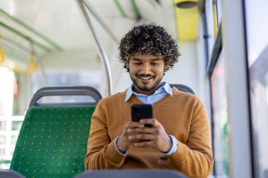 Otobüste oturan genç adam akıllı telefon kullanıyor, gülümsüyor ve dijital içerikle meşgul. Günlük kıyafet ve rahat atmosfer, iş çıkışı sırasında modern kentsel yaşam tarzını ve bağlantıyı vurguluyor.