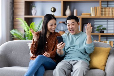 Asyalı bir aile evdeki kanepede otururken telefonda heyecan verici haberlerin tadını çıkarıyor. Olgun çift mutluluk ve şaşkınlık ifade ediyor, teknoloji ve iletişimi samimi bir ortamda vurguluyor.