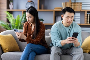 Kanepede oturan Asyalı çift, her biri telefon kullanıyor. Her ikisi de teknolojinin modern ilişkilerdeki rolünü gösteren ekranlara odaklanmış görünüyor. Dijital iletişim ve bağlantı anını yakalar.