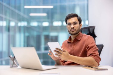 Modern ofis ortamında elinde tablet bulunan ciddi bir programcı dizüstü bilgisayarla dijital proje üzerinde çalışıyor. Akıllı günlük kıyafetler içinde iş profesyoneli, iş görevlerine odaklanıyor.