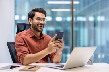 Ofiste oturan gülümseyen profesyonel bilgisayar ve defterler yakınlardayken akıllı telefon kullanıyor. Gözlük takmak ve günlük giysiler giymek, olumlu ifade gösterir, bağlanabilirliği gösterir.