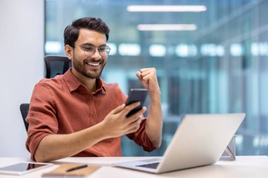 Gündelik gömlekli genç adam ofis masasında akıllı telefon kullanırken neşesini ifade ediyor. Dizüstü bilgisayar açık, mutlu ve muzaffer görünüyor, bu da olumlu başarıyı gösteriyor. Başarının atmosferi