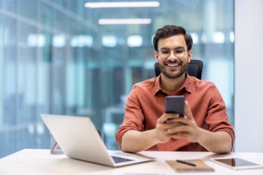 Ofis masasında oturmuş gülümseyen adam modern kurumsal ortamda iş iletişimi için akıllı telefonu kullanıyor. Bilgisayar ve telefon teknolojisiyle uzaktan çalışma kolaylığının keyfini çıkaran kişi.