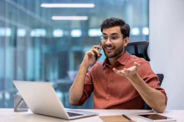Ofisteki kendine güveni tam bir adam dizüstü bilgisayar kullanırken telefonla konuşuyor. Teknoloji, iletişim, iş profesyonelliği unsurlarını gösteriyor. Modern çalışma ortamının özünü yakalar