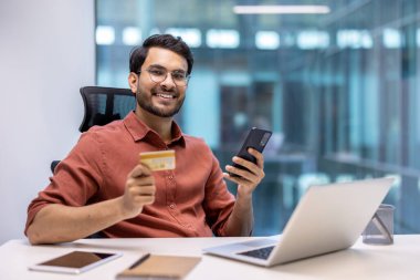 Ofiste elinde telefon ve kredi kartı olan gülümseyen bir adam. Çevrimiçi bankacılık, dijital ödeme, iş ve teknoloji kavramı. Dijital olarak bağlanan kendinden emin ve mutlu profesyonel.