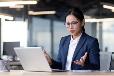 Asyalı iş kadını dizüstü bilgisayarından kötü haberler alıyor, şaşırıyor ve endişeleniyor. Modern ofis ayarları profesyonel ortamı vurgular.