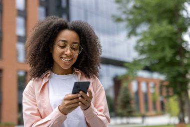 Kıvırcık saçlı neşeli bir kadın telefonuyla modern bir ofis binasının önünde güneşli günün tadını çıkarıyor..
