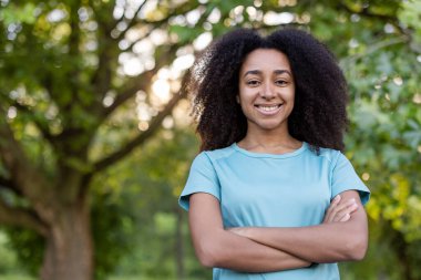 Kıvırcık saçlı genç bir kadın, kolları çapraz bir parkta, mavi giyinmiş, güneşli bir günü ve pozitifliği yakalıyor..