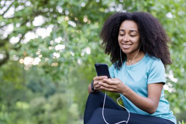 Gülümseyen genç bir kadın açık havada akıllı telefonuyla müzik dinliyor yemyeşil bir ortamda huzurlu bir günün tadını çıkarıyor..