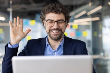 Takım elbiseli ve gözlüklü gülümseyen işadamı bir video görüşmesine katılıyor. Bir dizüstü bilgisayar kullanarak çağdaş bir ofis alanından sanal iş iletişimi sağlandı.