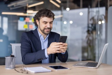 Takım elbiseli bir işadamı, bilgisayarla ve tabletle çevrili, modern bir ofiste akıllı telefon kullanırken gülümsüyor, dijital iletişimi ve birden fazla görevi gösteriyor..