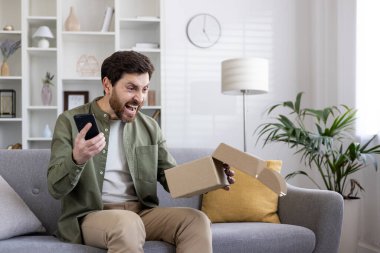 Kızgın erkek alıcı, elinde telefon, elinde karton kutuyla oturma odasındaki kanepede oturan hasarlı paketi aldı..