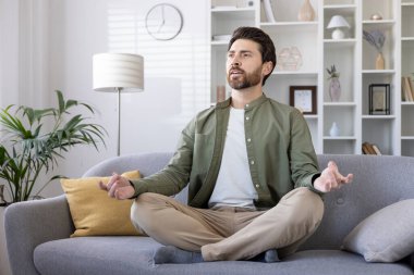 Modern oturma odasındaki kanepede meditasyon yapan adam. Huzurlu ev ortamında farkındalığı ve rahatlığı kucaklamak. Bağdaş kurmuş, gözleri kapalı, iç huzur ve sükunete odaklanmış biri..