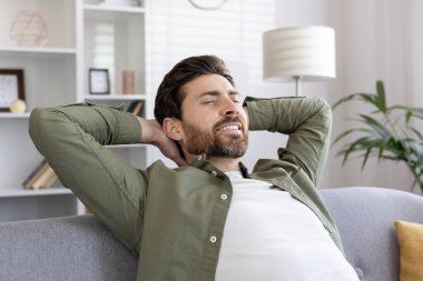 Rahat ve rahat bir kanepe deneyimi yaşayan sıradan giyinmiş bir adam. Ellerini başının arkasına koy, huzur ve mutluluk göster. Parlak ve havadar oturma odası ortamı memnuniyet hissi veriyor..