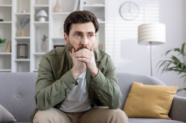 Gündelik giysiler içinde düşünceli bir adam modern oturma odasındaki kanepede oturur, düşüncelere dalmış bir şekilde. Sakin atmosfer, içgözlem ve seçim yapma anlamına gelir. Duygu aktarımı düşünceli, düşünceli bir yansımadır.