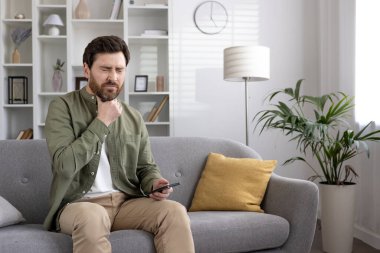 Gri kanepede oturan bir adam boğaz rahatsızlığı yaşıyor, telefonda rahatsız edici bir ifadeyle konuşuyor. Sahne, sağlık endişesi, hastalık ve modern ev ortamında rahatlama kavramını yakalıyor.