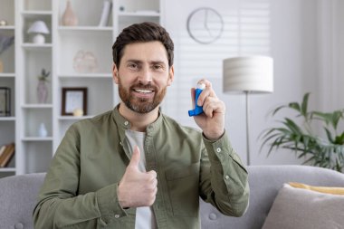 Astım spreyini kullanan kendine güvenen bir adam başparmaklarını kaldırırken, pozitifliği iletirken ve solunum hastalığının etkili bir şekilde yönetilmesini sağlarken. Kapalı mekanlar, sağlık farkındalığının ve yaşam tarzının geliştirilmesi.