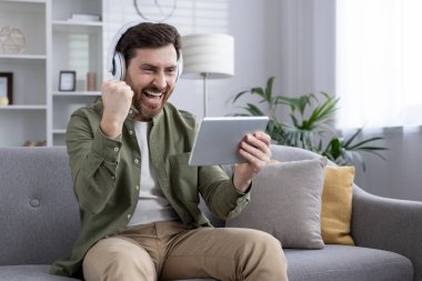 Kulaklık takan bir adam tabletteki dijital içerikle uğraşırken elini kaldırarak heyecanını dile getiriyor. Gelişigüzel taşıma, rahatlama, keyif, başarı..