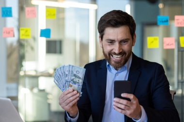 İş adamı elinde para tutarak ve akıllı telefonu kontrol ederek mutluluğunu ifade ediyor. Resmi takım elbiseli adam, zenginlik, iletişim ve iş kavramlarını tasvir ederek başarı ve mali başarı gösteriyor..