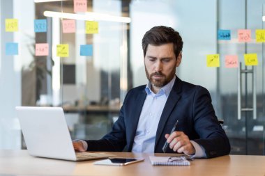 İşadamı dizüstü bilgisayar kullanarak ve notlar yazarak işine odaklanıyor. Profesyonel ortam odaklanmayı, üretkenliği ve modern çalışma ortamını vurgular. Ofis sahnesi planlama ve stratejik düşünme gösteriyor