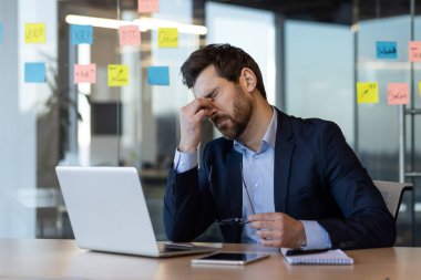 İş adamı kendini stresli ve bunalmış hissediyor, hayal kırıklığı içinde masada oturuyor. Laptop, notlar ve düşünceli ifade ile ofis ortamı iş baskısı ve zihinsel yorgunluğu iletir.